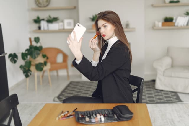 Young woman in stylish outfit holding master class for makeup in internet while sitting on chair at table with set of cosmetics and brushes in light living room